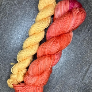 Könnte beinhalten: Zwei Stränge Garn in Gelb- und Orangetönen. Das gelbe Garn ist einfarbig, während das orangefarbene Garn einen Farbverlauf aufweist, der von Rosa zu Orange übergeht. Das Garn ist zu einer seilartigen Form verdreht.