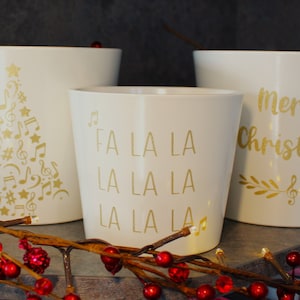 May include: Three white ceramic plant pots with gold festive designs. One features a musical Christmas tree, another says "Fa La La La," and the third reads "Merry Christmas."
