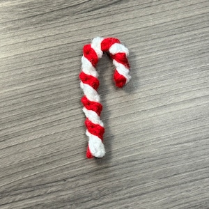 May include: A red and white candy cane made of yarn, on a woodgrain background.