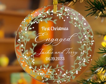 May include: Clear glass Christmas ornament with a white wreath design and the text "First Christmas Engaged Jackie and Terry 06.19.2023"