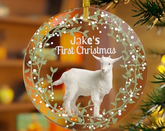 Könnte beinhalten: Ein runder Weihnachtsbaumschmuck aus Glas mit einer weißen Ziege und grünen Blättern. Der Schmuck ist mit dem Text "Jake's First Christmas" verziert.