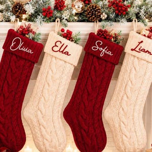 Puede incluir: Cuatro calcetines navideños de punto trenzado colgados en fila, alternando entre los colores rojo oscuro y crema. Cada calcetín presenta un puño doblado bordado con los nombres Olivia, Ella, Sofia y Liam en letra cursiva.