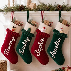 Op de afbeelding: Vier kerstsokken van afwisselend rood en groen fluweel met witte boorden en ruches. Elke sok is voorzien van wit geborduurde namen: Mommy, Daddy, Sophie en Amelia. Decoratieve viltapplicaties tonen een rendier, boom, kerstman en sneeuwpop.