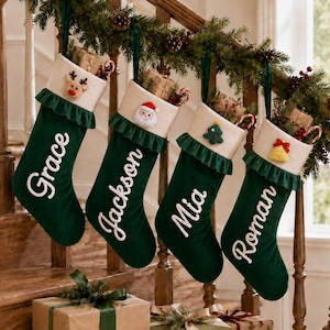 May include: Four dark green Christmas stockings with white cuffs and ruffled trim. Each stocking features a white embroidered name: Grace, Jackson, Mia, or Roman. Decorative felt appliques include a reindeer, Santa, tree, and bell.
