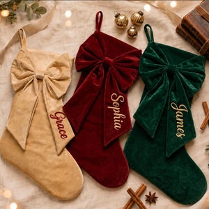 Puede incluir: Tres calcetines navideños de terciopelo en beige, rojo oscuro y verde oscuro, cada uno con un gran lazo decorativo en la parte superior. Los calcetines están personalizados con nombres bordados: Grace en rojo sobre el calcetín beige, y Sophia y James en dorado sobre los calcetines rojo y verde.