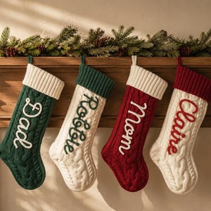 May include: Four cable-knit Christmas stockings hanging in a row. The first is dark green with a white cuff and the name Dad. The second is white with a dark green cuff and the name Robbie. The third is dark red with a white cuff and the name Mom. The fourth is white with a dark red cuff and the name Claire.