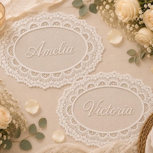 May include: Two oval-shaped white lace doilies featuring intricate scalloped edges. The top doily displays the name Amelia in white script lettering, while the bottom doily features the name Victoria in matching white script lettering.