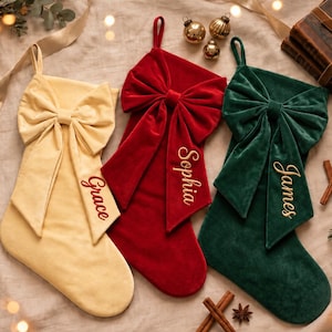 May include: Three velvet Christmas stockings in cream, red, and dark green, each featuring a large decorative bow at the top. The stockings are personalized with embroidered names: Grace in red, Sophia in gold, and James in gold thread.