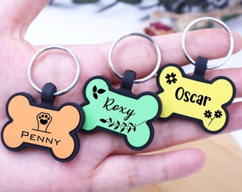 May include: Three dog bone-shaped keychains with different colors and names. The first keychain is orange with the name "PENNY" and a paw print. The second keychain is green with the name "ROXY" and a floral design. The third keychain is yellow with the name "OSCAR" and a floral design.
