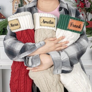 May include: Three cable-knit Christmas stockings in red, beige, and white, each with a personalized wooden name tag. The stockings are held by a person in a plaid shirt, with festive decorations in the background.