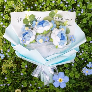 Könnte beinhalten: Ein Strauß handgefertigter blau-weißer Häkelblumen, verpackt in hellblauem Papier und mit einem Band gebunden. Der Strauß enthält eine Karte mit den Worten "You are sunshine". Der Strauß befindet sich vor einem Hintergrund aus grünem Laub.