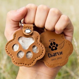 May include: Two brown leather paw-shaped keychains. One keychain has a clear window with white fur inside. The other keychain has a paw print with angel wings, the name "Charlie", and the years 2012-2023.
