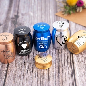 Może przedstawiać: Zestaw małych, grawerowanych metalowych urn w różnych kolorach, w tym złoty, srebrny, czarny i niebieski. Każda urna zawiera spersonalizowany napis i wzór, taki jak serce, motyl lub gołąb. Urny są ułożone na drewnianej powierzchni.