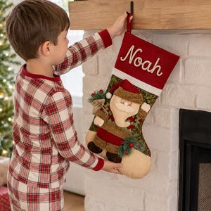 Puede incluir: Un calcetín navideño con un puño rojo que tiene el nombre Noah bordado en letra blanca. El cuerpo del calcetín muestra una aplicación texturizada de Santa Claus con un suéter verde estampado, sosteniendo un pequeño saco y una rama de acebo.