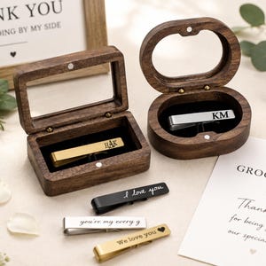 May include: Two dark wood boxes with glass lids, one rectangular and one oval, each containing a metal tie bar engraved with initials. Three additional tie bars are displayed outside the boxes, featuring engraved phrases like I love you.