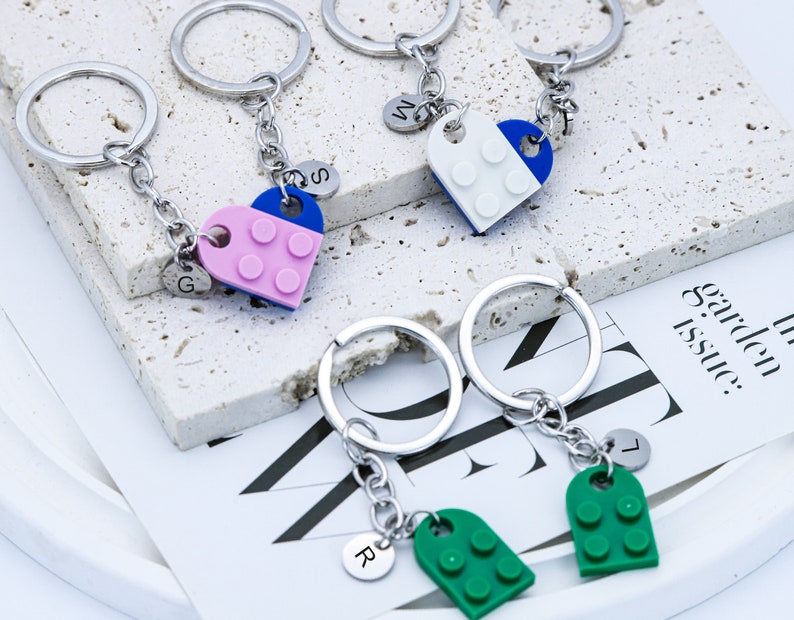 May include: Set of four silver keychains with heart-shaped charms. Each charm is made of plastic and features a different color combination of blue, white, pink, and green. The charms are shaped like Lego bricks and have the letters G, S, W, and L on the back.