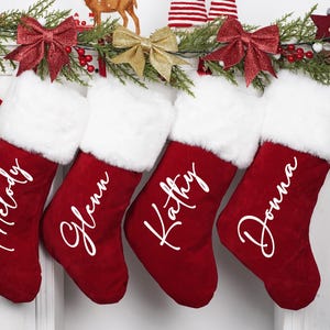 May include: Four red Christmas stockings with white faux fur trim and personalised names in white script: Melody, Glenn, Kathy, and Donna. The stockings are decorated with bows and greenery.