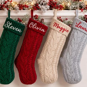 May include: Four cable-knit Christmas stockings in dark green, red, cream, and light grey. Each stocking features a ribbed cuff embroidered with a name in white or red script: Steven, Olivia, Maggie, and Lauren. The stockings are hung in a row.