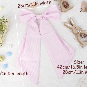 May include: A pink and white striped bow, 42cm long and 28cm wide, lies on a white surface. Decorative wooden Easter bunnies and flowers surround the bow. The bow is a hair accessory.