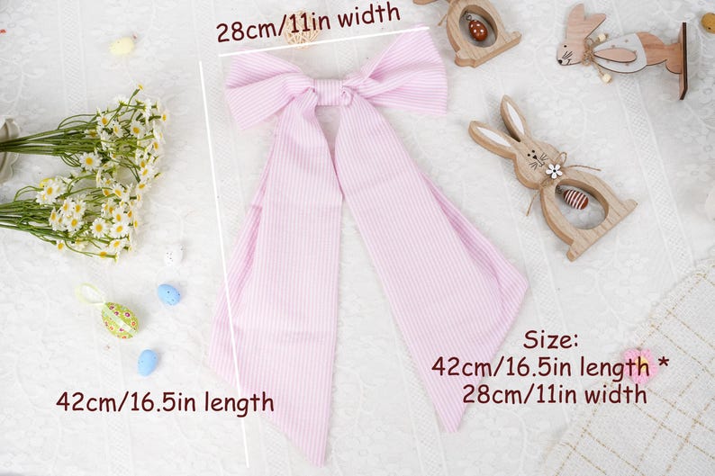 May include: A pink and white striped bow, 42cm long and 28cm wide, lies on a white surface. Decorative wooden Easter bunnies and flowers surround the bow. The bow is a hair accessory.