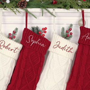 Op de afbeelding: Vier gepersonaliseerde kerstsokken, twee rode en twee witte, hangen aan een witte schoorsteenmantel. Elke sok is geborduurd met een naam in een complementaire kleur. Groen en bessen sieren de bovenkant van elke sok.
