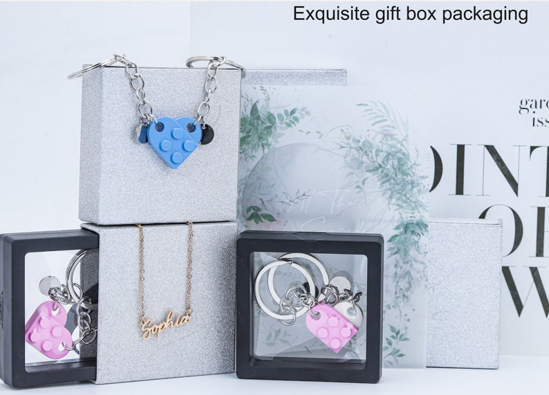 May include: Three gift boxes with jewelry inside. The top box has a silver chain necklace with a blue heart-shaped charm. The middle box has a silver chain necklace with a gold nameplate that says "Sophia". The bottom box has a silver keychain with a pink heart-shaped charm.