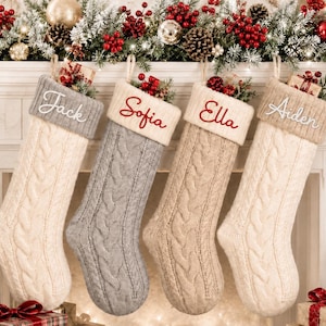 Puede incluir: Cuatro calcetines navideños de punto trenzado colgados en fila. El primero es crema con un puño gris bordado con 'Jack'. El segundo es gris con un puño crema bordado con 'Sofia'. El tercero es beige con un puño crema bordado con 'Ella'. El cuarto es crema con un puño beige bordado con 'Aiden'.