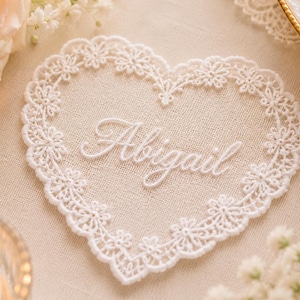 Può includere: Un centrino in pizzo bianco a forma di cuore con un intricato motivo floreale sul bordo e il nome Abigail ricamato in corsivo bianco al centro. Il pizzo è realizzato con un delicato design traforato e una finitura smerlata.