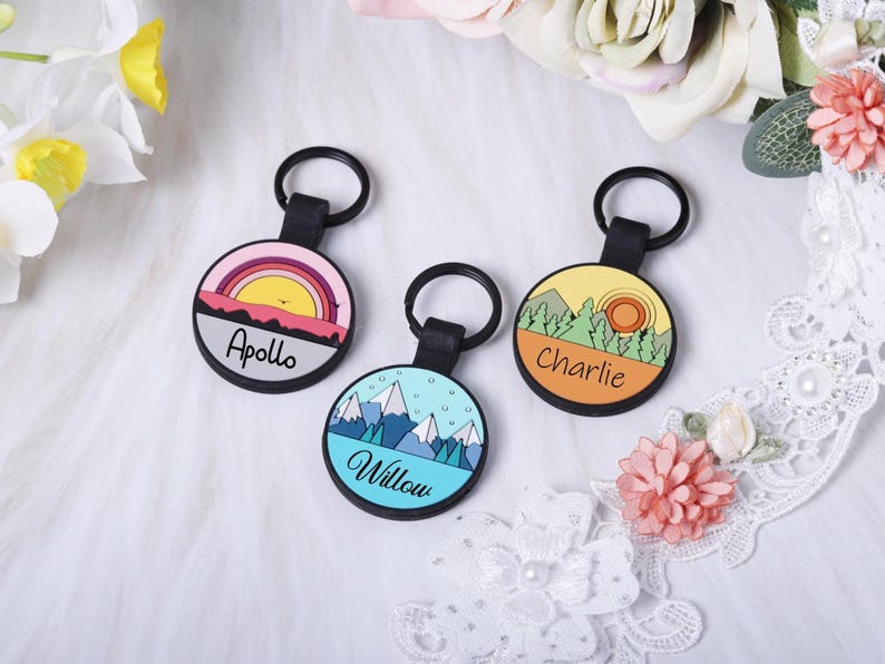 Puede incluir: Tres llaveros redondos con anillos negros. Cada llavero presenta un dise&ntilde;o de paisaje diferente con un nombre: "Apollo" con una puesta de sol, "Willow" con monta&ntilde;as y "Charlie" con &aacute;rboles y un sol. Los llaveros tienen correas de cuero negro.
