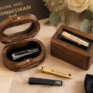 May include: Two dark wood boxes, one oval and one rectangular, containing metal tie clips. One silver clip is engraved with 'Phelps', one gold clip with 'A Father's Love Learned', one gold with 'We love you' and a heart, and one black with 'HB'.