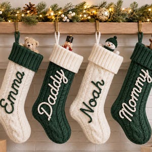 Op de afbeelding: Vier kerstsokken van kabelgebreid materiaal hangen op een rij, afwisselend in de kleuren crème en donkergroen. Elke sok is voorzien van een contrasterende geborduurde naam: Emma, Daddy, Noah en Mommy. Kleine knuffels steken boven de rand uit.