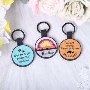 Puede incluir: Tres etiquetas redondas de identificaci&oacute;n para mascotas con llaveros negros. Una etiqueta es de color azul claro con huellas de patas y el texto "CALL MY DADDY". Otra etiqueta tiene un dise&ntilde;o de puesta de sol y el nombre "Barker".