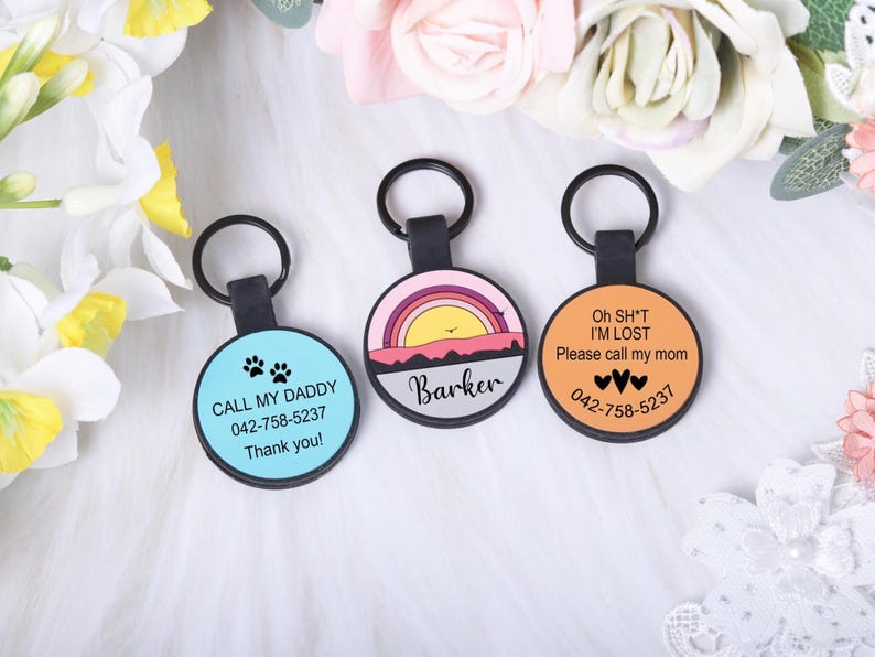 Puede incluir: Tres etiquetas redondas de identificaci&oacute;n para mascotas con llaveros negros. Una etiqueta es de color azul claro con huellas de patas y el texto "CALL MY DADDY". Otra etiqueta tiene un dise&ntilde;o de puesta de sol y el nombre "Barker".