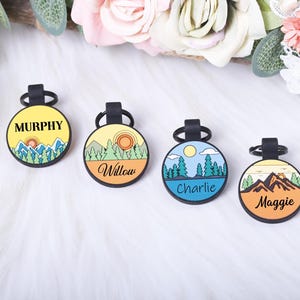 Puede incluir: Cuatro etiquetas redondas para mascotas con lazos negros. Cada etiqueta presenta un dise&ntilde;o de paisaje y un nombre: Murphy, Willow, Charlie y Maggie. Las etiquetas tienen un esquema de color amarillo, naranja y azul, con ilustraciones de monta&ntilde;as y &aacute;rboles.