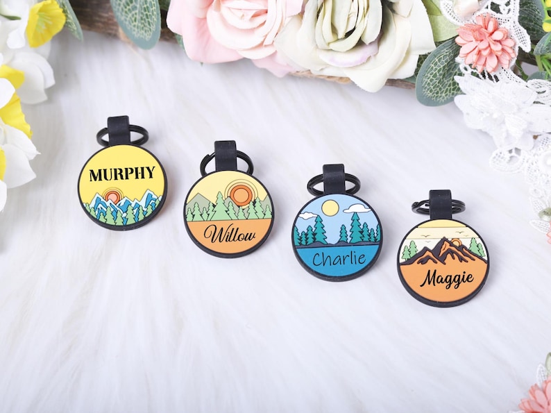 Puede incluir: Cuatro etiquetas redondas para mascotas con lazos negros. Cada etiqueta presenta un dise&ntilde;o de paisaje y un nombre: Murphy, Willow, Charlie y Maggie. Las etiquetas tienen un esquema de color amarillo, naranja y azul, con ilustraciones de monta&ntilde;as y &aacute;rboles.