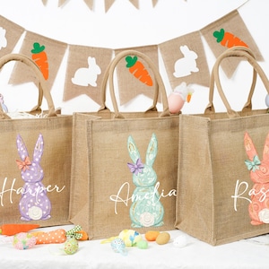 May include: Three hessian tote bags with bunny designs and names: Harper, Amelia, and Rascal. Each bag features a different coloured bunny with a bow. A banner with bunny and carrot cutouts hangs in the background. Easter-themed.