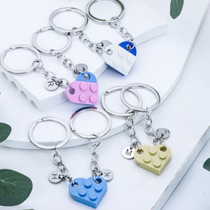 May include: Set of four heart-shaped keychains made of plastic building blocks. Each keychain has a different color: blue, pink, yellow, and white. Each keychain has a letter charm attached to it: R, H, K, and M.