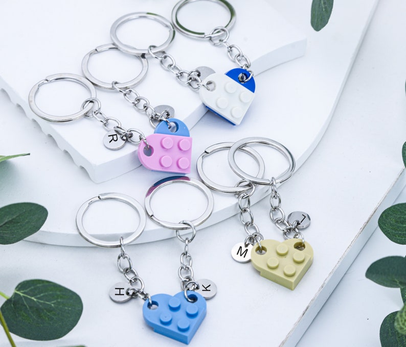 May include: Set of four heart-shaped keychains made of plastic building blocks. Each keychain has a different color: blue, pink, yellow, and white. Each keychain has a letter charm attached to it: R, H, K, and M.