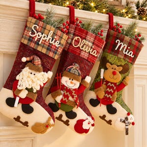 Könnte beinhalten: Drei Weihnachtsstrümpfe mit karierten Bündchen, auf denen die Namen Sophie, Olivia und Mia in Weiß gestickt sind. Die Strümpfe zeigen Plüschfiguren von Weihnachtsmann, Schneemann und Rentier, verziert mit roten Beeren und Tannenzweigen.