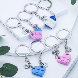 May include: Five silver keychains with heart-shaped charms. Each charm is made of plastic and resembles a Lego brick. The charms are blue, pink, and white. The keychains have letter charms attached to them, spelling out the letters "K", "L", "D", "U", and "J".