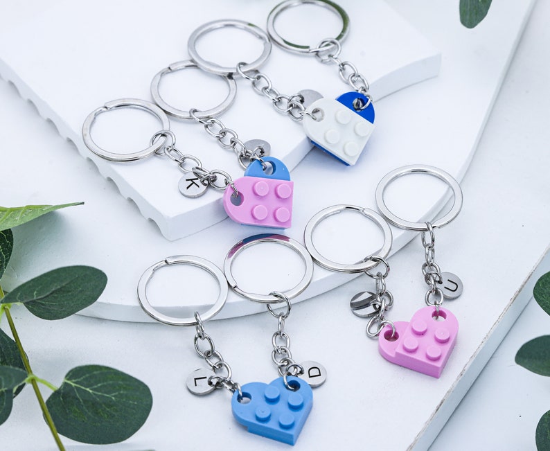 May include: Five silver keychains with heart-shaped charms. Each charm is made of plastic and resembles a Lego brick. The charms are blue, pink, and white. The keychains have letter charms attached to them, spelling out the letters "K", "L", "D", "U", and "J".
