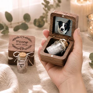 K&ouml;nnte beinhalten: Eine dunkle Holz-Erinnerungsbox mit Klappdeckel, die ein kleines Glasfl&auml;schchen enth&auml;lt. Die Innenseite des Deckels zeigt ein Foto eines schwarz-wei&szlig;en Hundes. Eine passende geschlossene Box tr&auml;gt die Gravur Penny 2012-2023 mit Pfotenabdruck.