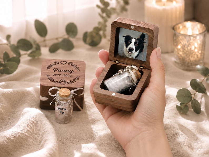 K&ouml;nnte beinhalten: Eine dunkle Holz-Erinnerungsbox mit Klappdeckel, die ein kleines Glasfl&auml;schchen enth&auml;lt. Die Innenseite des Deckels zeigt ein Foto eines schwarz-wei&szlig;en Hundes. Eine passende geschlossene Box tr&auml;gt die Gravur Penny 2012-2023 mit Pfotenabdruck.