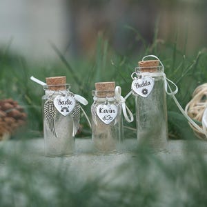May include: Three small glass bottles with cork tops and white ribbon. Each bottle contains white feathers and a silver charm with a name on it. The names are Sadie, Kevin, and Hedda.