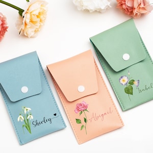 Könnte beinhalten: Drei personalisierte Beutel mit Blumendesigns und Namen. Die Beutel sind hellblau, pfirsichfarben und grün. Der blaue Beutel hat ein Schneeglöckchen-Design und den Namen "Shirley". Der pfirsichfarbene Beutel hat ein rosafarbenes Rosen-Design und den Namen "Abigail". Der grüne Beutel hat ein Winden-Design und den Namen "Isabella".