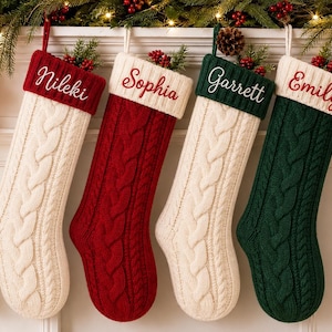 Puede incluir: Cuatro calcetines de Navidad personalizados con un patrón de punto trenzado. Dos calcetines son crema con puños rojos bordados con los nombres Nileki y Sophia, y dos son rojos y verdes con puños crema bordados con los nombres Garrett y Emily.