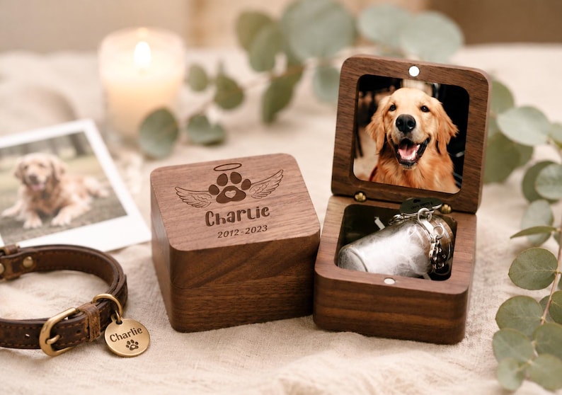 Pode incluir: Uma caixa quadrada de madeira escura com tampa articulada contendo a foto de um golden retriever. A tampa externa &eacute; gravada com uma pata, asas de anjo, uma aur&eacute;ola e o texto Charlie 2012-2023. Um pequeno frasco de vidro est&aacute; dentro da caixa.