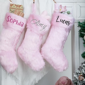 May include: Three pink Christmas stockings, each with a name embroidered on the cuff: "Sophia," "Amy," and "Lucas." The stockings are made of a soft, furry material. A small Christmas tree and decorations are visible in the background.