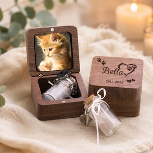 Pode incluir: Uma caixa de recorda&ccedil;&atilde;o de madeira escura com tampa articulada apresentando a foto de um gatinho laranja. A tampa est&aacute; gravada com o nome Bella, pegadas, um cora&ccedil;&atilde;o e os anos 2012-2023. Um pequeno frasco de vidro com rolha est&aacute; dentro.