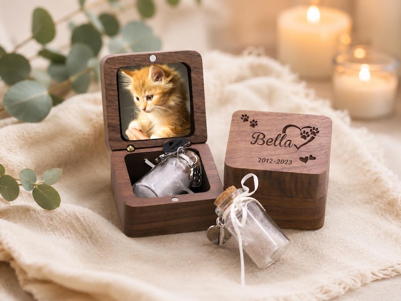 Pode incluir: Uma caixa de recorda&ccedil;&atilde;o de madeira escura com tampa articulada apresentando a foto de um gatinho laranja. A tampa est&aacute; gravada com o nome Bella, pegadas, um cora&ccedil;&atilde;o e os anos 2012-2023. Um pequeno frasco de vidro com rolha est&aacute; dentro.